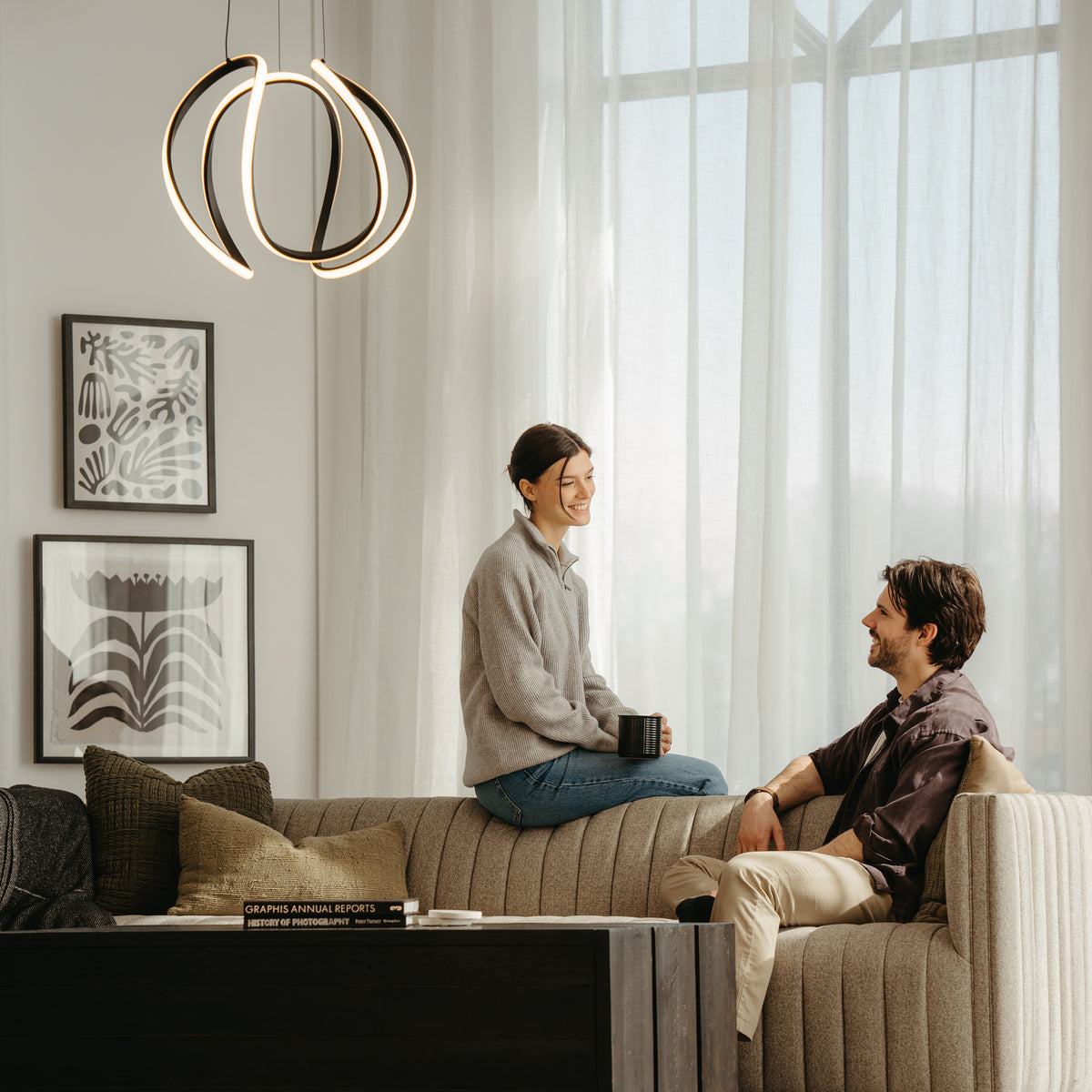 A woman sits on the back of a beige sofa holding a mug, smiling at a man seated on the sofa. Modern decor, framed abstract art, and a unique hanging light fixture are visible in the cozy, sunlit living room.