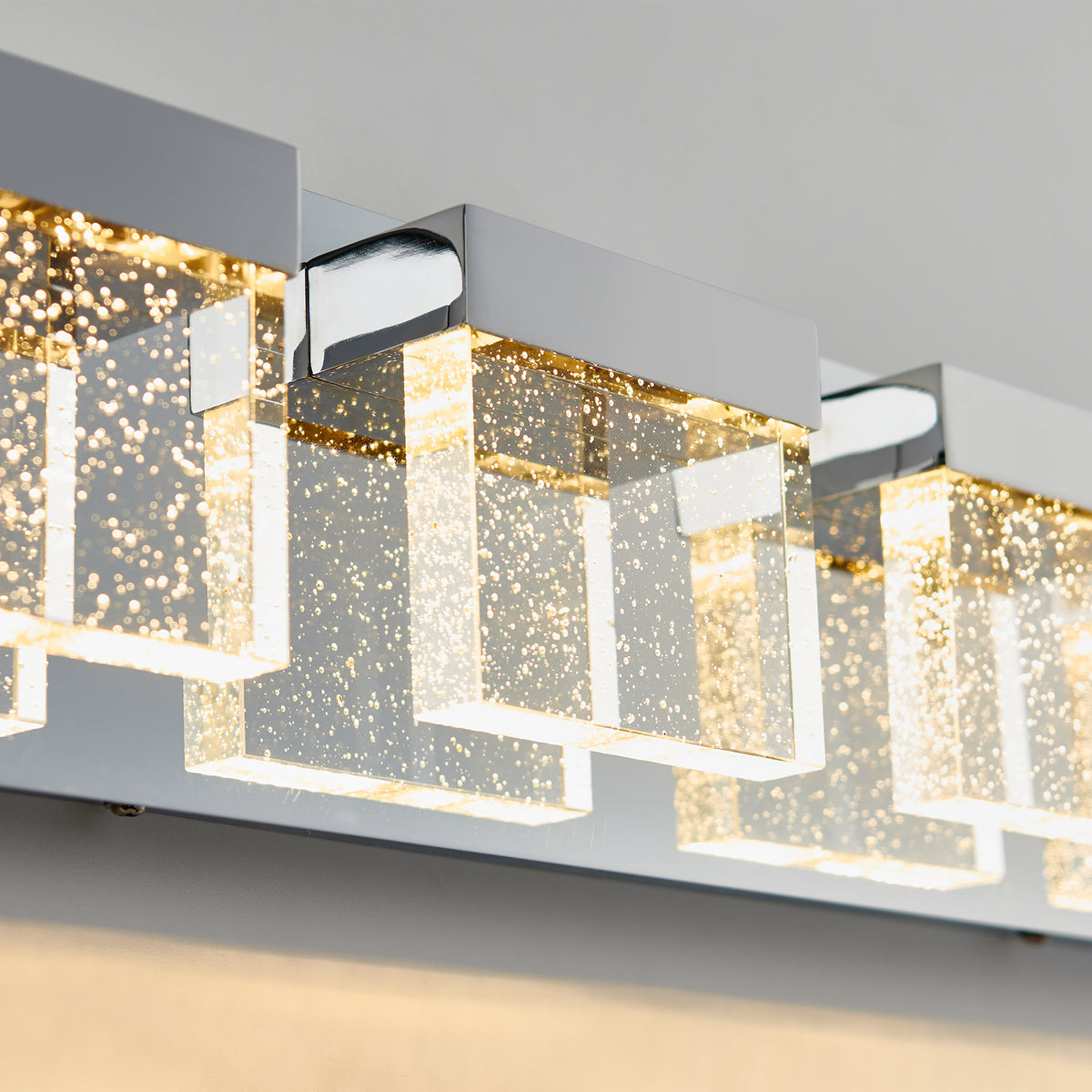 Close-up of a modern wall light fixture with rectangular glass shades containing small bubbles, mounted on a shiny chrome base and emitting a warm, glowing light.