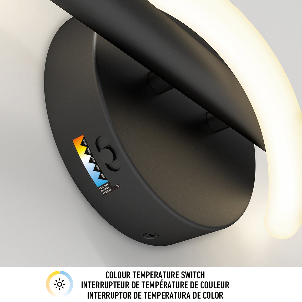 Close-up of a modern black wall light fixture with a curved illuminated arm. A colourful label and a colour temperature switch symbol are visible on the fixture base. Text below describes a colour temperature switch feature in three languages.