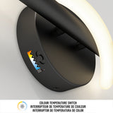 Close-up of a modern black wall light fixture with a curved illuminated arm. A colourful label and a colour temperature switch symbol are visible on the fixture base. Text below describes a colour temperature switch feature in three languages.