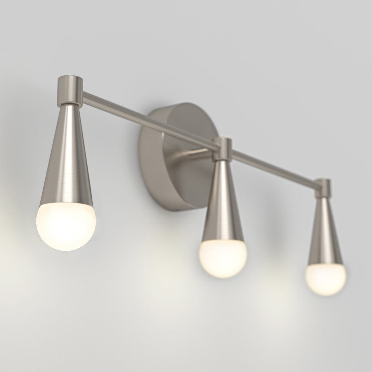 A modern wall-mounted light fixture with three brushed metal cone-shaped lamps, each with a glowing round bulb, arranged horizontally on a light-colored wall.