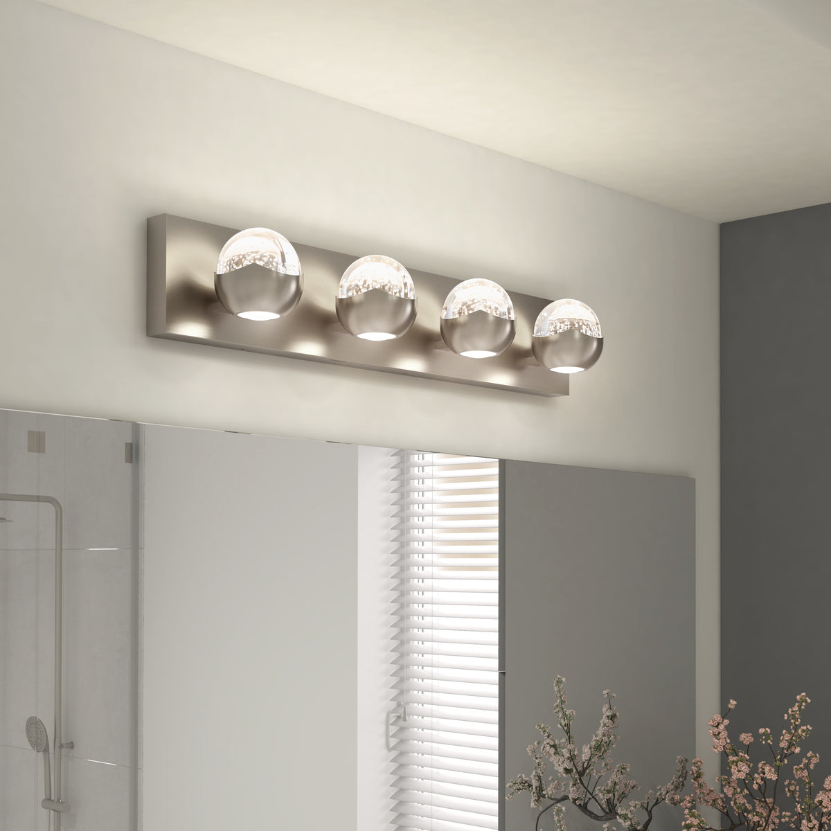 A modern bathroom light fixture with four spherical glass bulbs on a rectangular metal base is mounted above a mirror, with a window and decorative plants visible in the background.