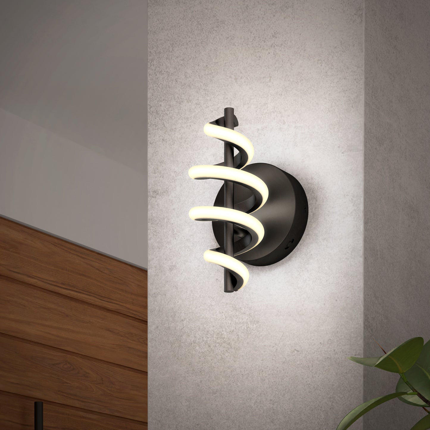 The Artika Chorus LED Wall Light, featuring a black circular base and spiral LED design, is mounted on a light gray wall with part of a wooden cabinet and green plant leaves visible nearby.