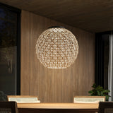 A round woven pendant light hangs above a wooden dining table with chairs, set in a room with wooden walls, a large window, and a plant in the corner.