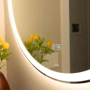 The Artika Mateo 28 in. LED mirror with 3 CCT and defogger is illuminated, showing yellow flowers in a vase reflected in its anti-fog surface, with a wall and framed photo visible in the background. Touch buttons control power and defog functions.