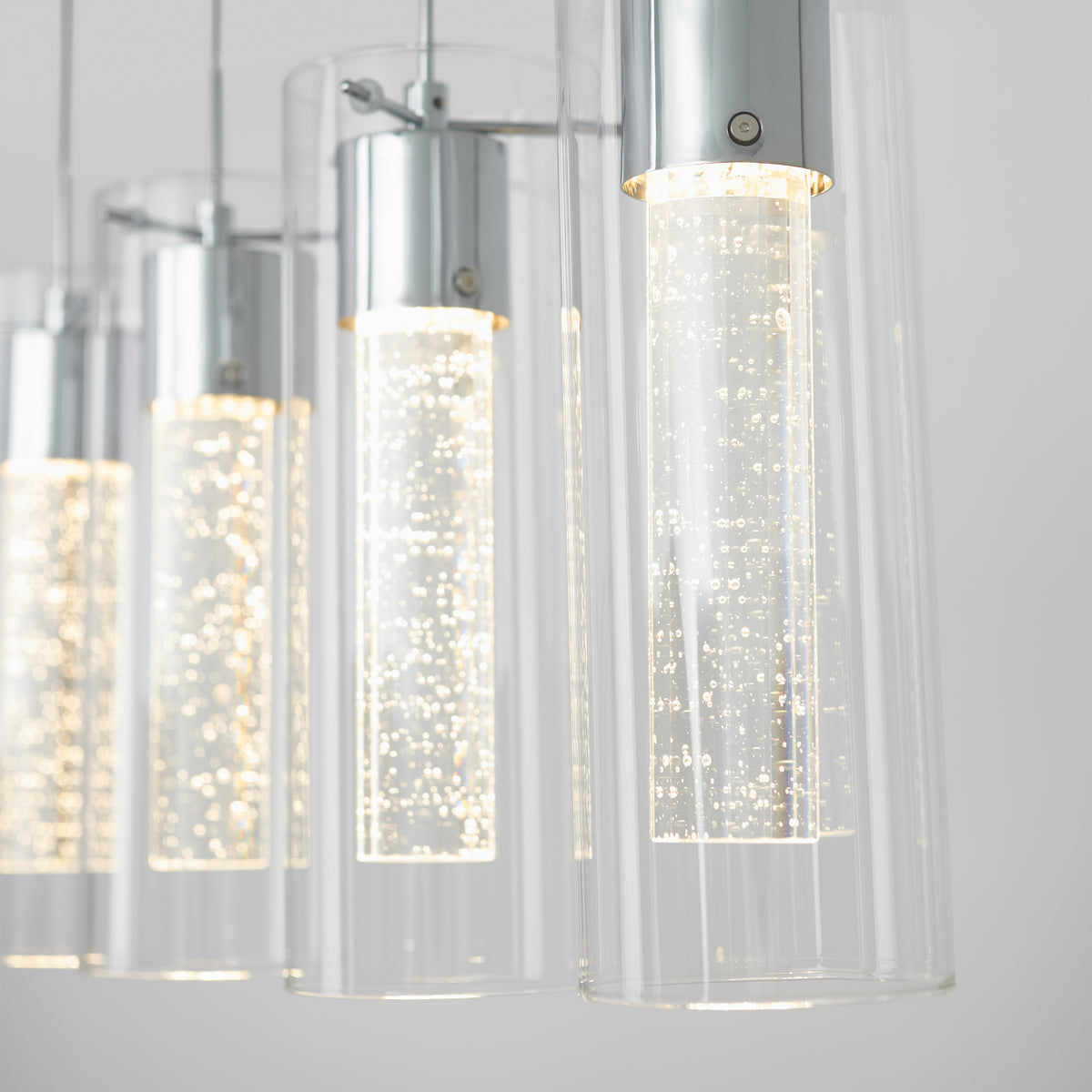 Close-up of modern pendant lights with cylindrical glass shades and chrome fittings, featuring inner tubes with tiny bubbles illuminated by bright LED bulbs against a light background.