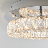 Close-up view of a modern ceiling light fixture with a circular design, featuring clear, faceted crystal elements that sparkle and reflect the light, mounted on a metallic base against a white background.