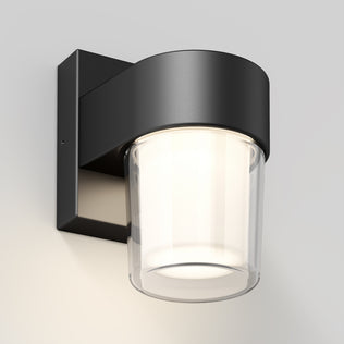 The Artika Halden indoor-outdoor LED wall light 5 CCT is a modern black wall-mounted fixture with a cylindrical frosted glass shade, casting a soft white glow on any light-colored wall.