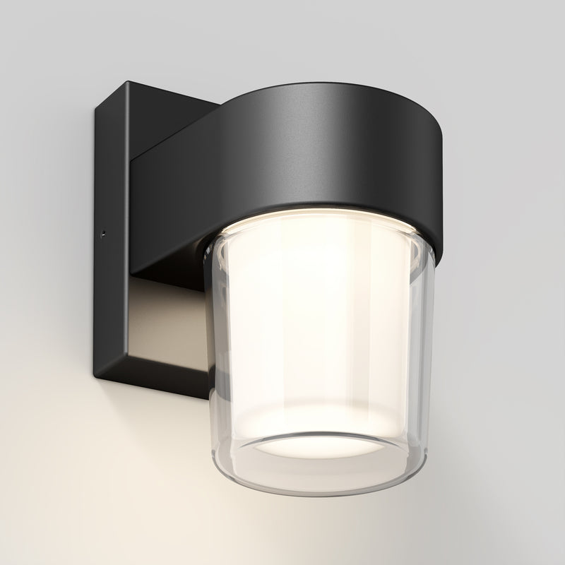 The Artika Halden indoor-outdoor LED wall light 5 CCT is a modern black wall-mounted fixture with a cylindrical frosted glass shade, casting a soft white glow on any light-colored wall.