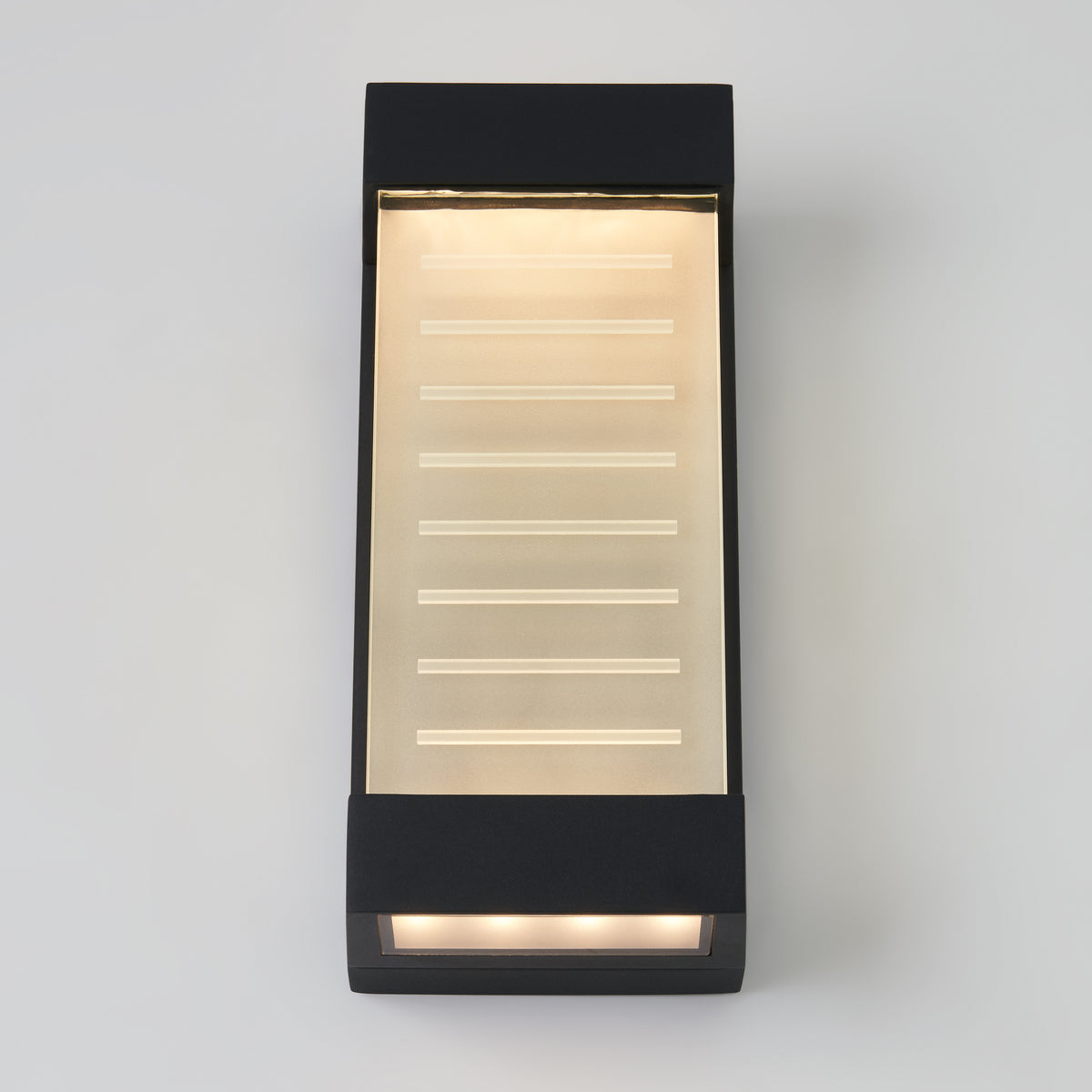 A modern rectangular black wall light fixture with a frosted glass panel, emitting a warm glow. Horizontal lines are etched on the glass, and light shines from both the top and bottom edges.
