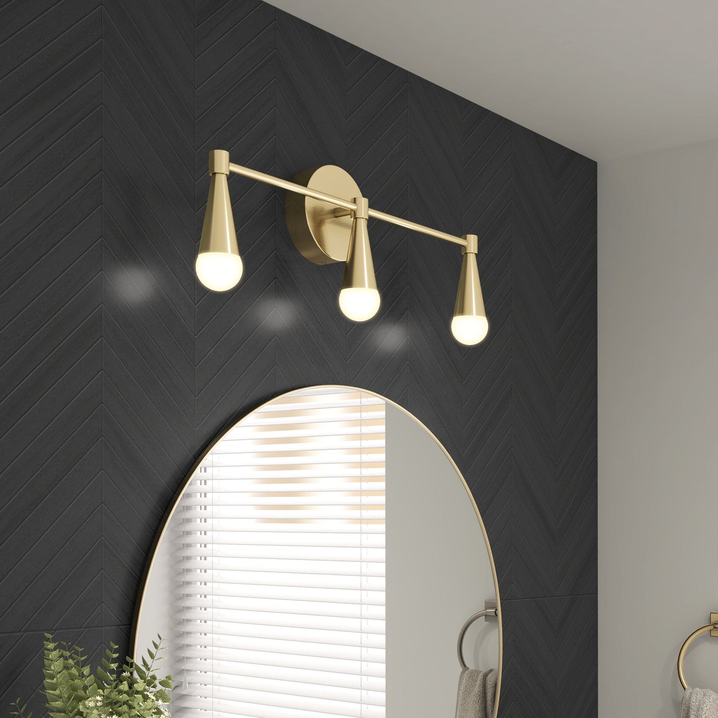 A modern bathroom features a round gold-framed mirror, a black herringbone tile wall, and an Artika Wayden LED mid-century bathroom vanity light (3 CCT) above the mirror. A towel ring and plant complete the look.