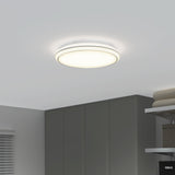 A modern, round ceiling light fixture is illuminated on a white ceiling above a minimalist room with gray cabinets, folded towels, and a window with blinds.