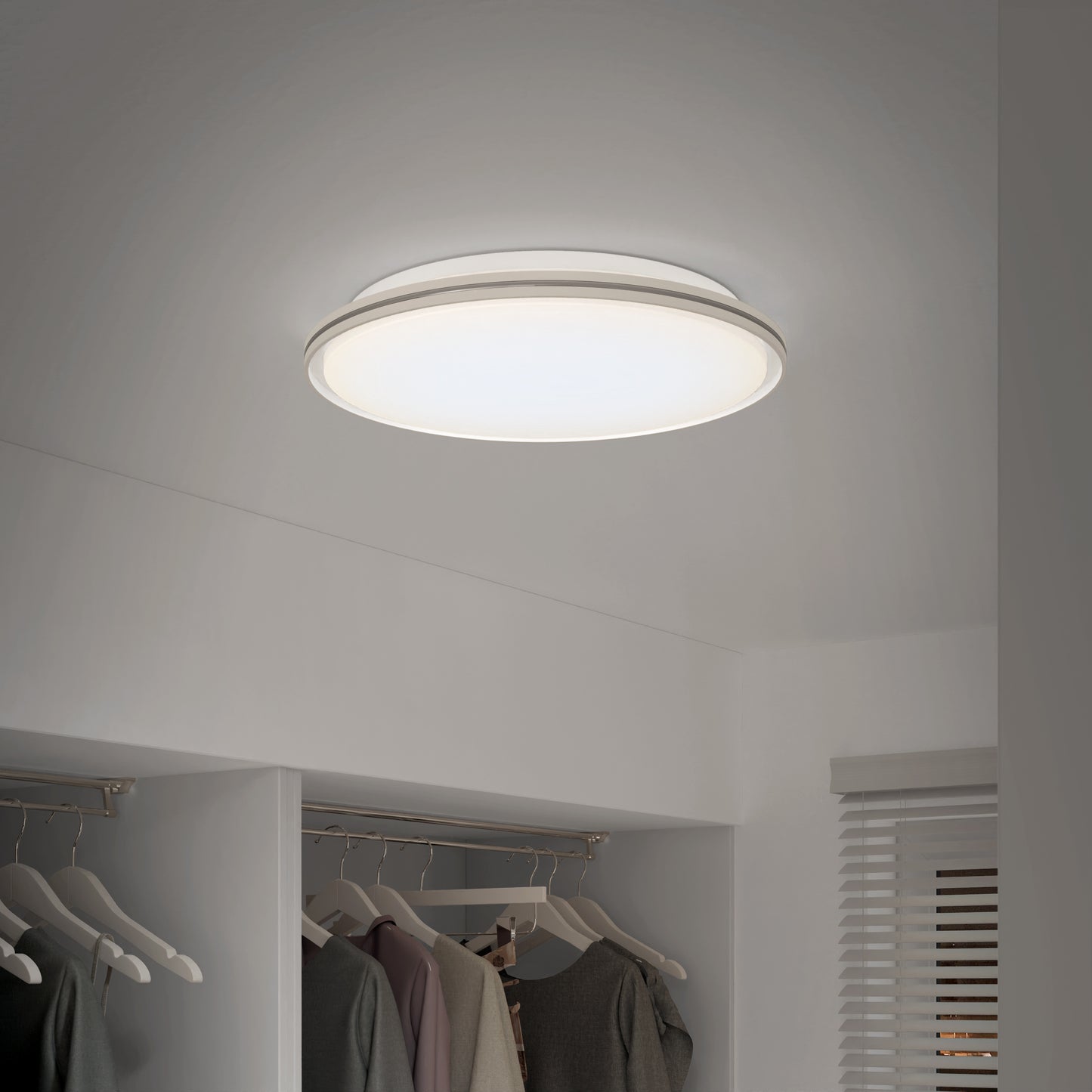 An Artika Astrid 16.5 in. LED flush mount light with 5 CCT and remote illuminates a tidy, minimalist white closet, where clothes hang below and a window with white blinds sits on the right wall.
