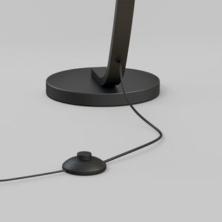 A close-up of the Artika Arc LED Floor Lamp base shows its power cord plugged in and a round foot switch, set against a light gray background.
