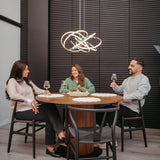 Three people sit around a round wooden dining table, smiling and holding wine glasses. They are in a modern room with dark textured walls and a decorative light fixture above them.