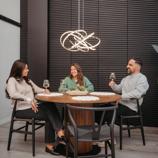 Three people sit around a round wooden table, smiling with wine glasses. Above them hangs the Artika Maelstrom modern LED pendant light 5 CCT, illuminating the space against a dark, textured wall.