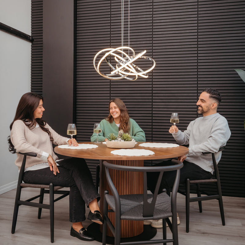 Three people sit around a round wooden table, smiling with wine glasses. Above them hangs the Artika Maelstrom modern LED pendant light 5 CCT, illuminating the space against a dark, textured wall.