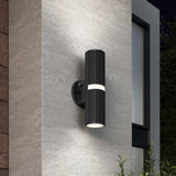 A modern black cylindrical outdoor wall light with horizontal grooves, mounted on a textured gray wall beside a window, illuminating upward and downward at night with plants visible below.