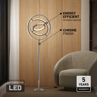 The Artika Pivot modern LED floor lamp 3 CCT with a sleek chrome finish and circular LED rings stands by a beige armchair and wood-paneled wall. Features include energy efficiency, integrated LED, and a 5-year warranty.