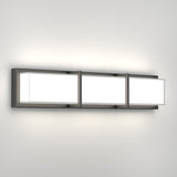 A modern wall-mounted light fixture with three rectangular, frosted glass panels set in a sleek, metallic frame, casting soft white light against a plain wall.