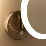 A close-up of a wall-mounted, circular makeup mirror with an illuminated LED light ring, featuring a metallic base and a button on the base, attached to a beige wall.