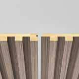 Two wooden panels with grooved, wavy surfaces and contrasting light wood edges are positioned side by side against a plain light background, with a small gap between them showing their interlocking design.