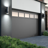 A modern gray garage door with rectangular windows at the top is framed by a white and dark exterior wall. Two cylindrical wall lights are illuminated on either side, and green shrubs line the foreground.