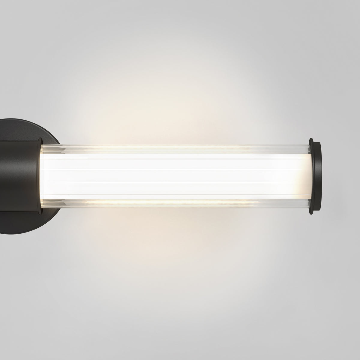 A modern cylindrical wall sconce with a black base and clear glass cover, emitting a soft white light against a light gray background.