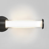 A modern cylindrical wall sconce with a black base and clear glass cover, emitting a soft white light against a light gray background.