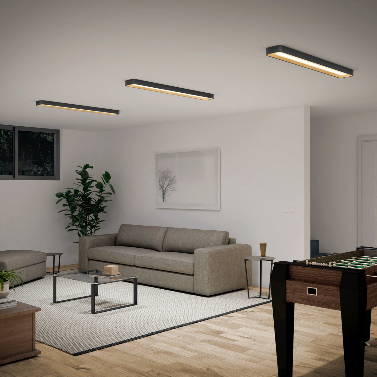 Modern living room with a gray sofa, glass coffee table, indoor plant, wall art, and foosball table. Three rectangular ceiling lights illuminate the space, which has wooden floors and neutral decor.