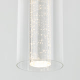A close-up of a modern cylindrical pendant light with clear glass and tiny bubbles inside, illuminated to create a sparkling effect. The background is plain and light-colored.