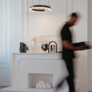 A blurred figure passes a modern white mantel adorned with books, a vase, globe, framed art, and candles. Overhead hangs the Artika Vance modern LED pendant light 3 CCT, illuminating the minimalist space.