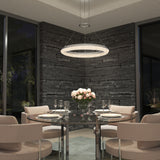 A modern dining room with a round glass table, beige chairs, a bouquet of pink roses, place settings, and glassware. Dark stone walls and large windows frame a glowing circular chandelier above.