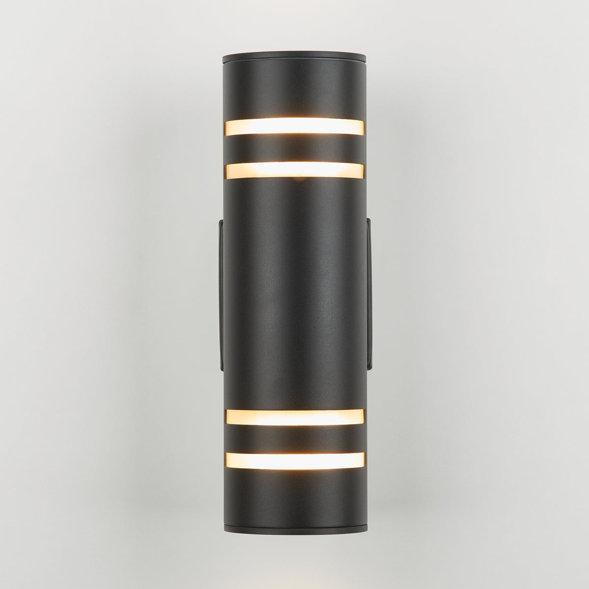 A modern cylindrical black wall sconce light fixture with horizontal frosted strips emits a soft white glow through openings near the top and bottom, set against a plain light gray background.