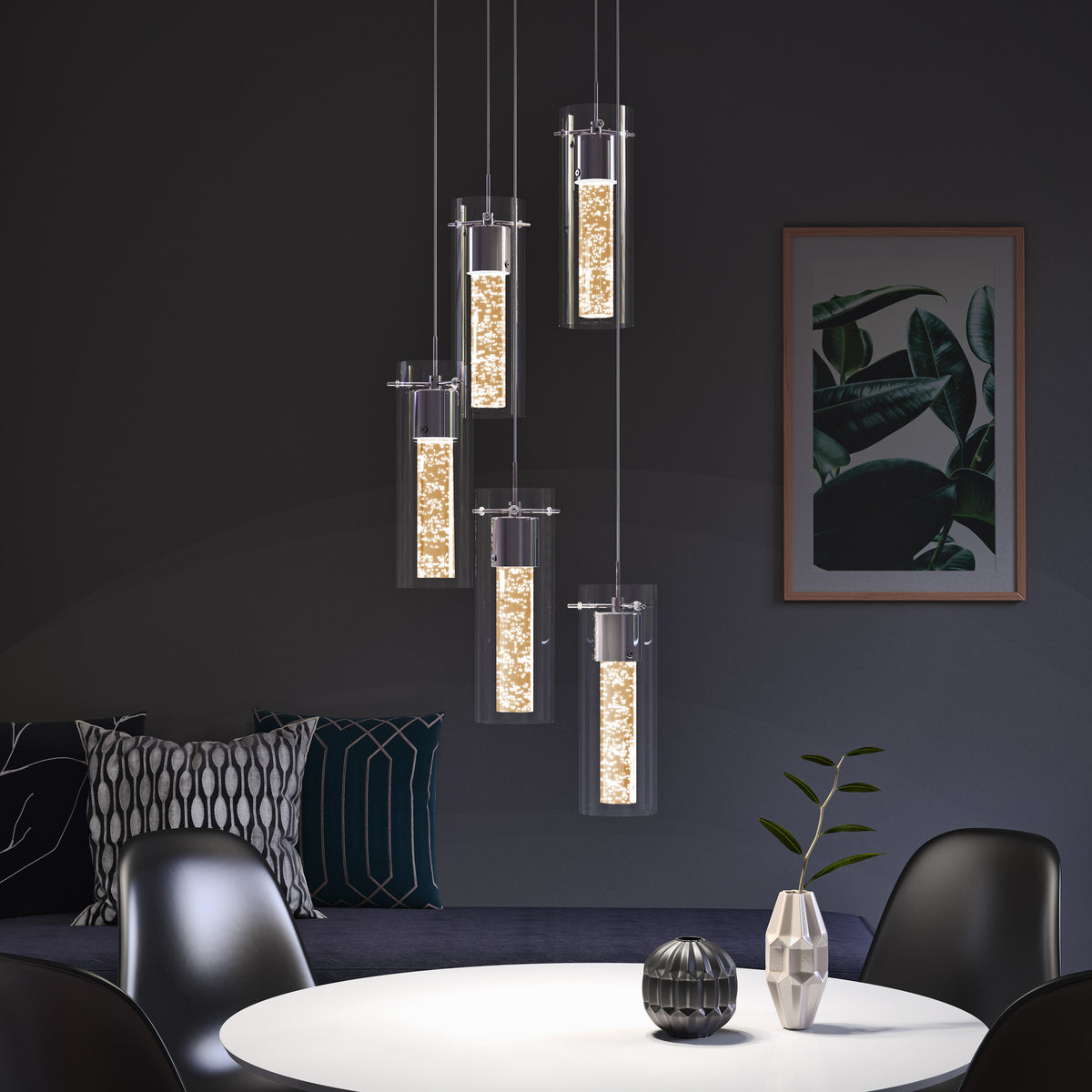 Modern dining room with a round white table, black chairs, a small vase with a branch, and six cylindrical pendant lights hanging above, glowing warmly. There are decorative pillows and a plant artwork on a dark wall in the background.