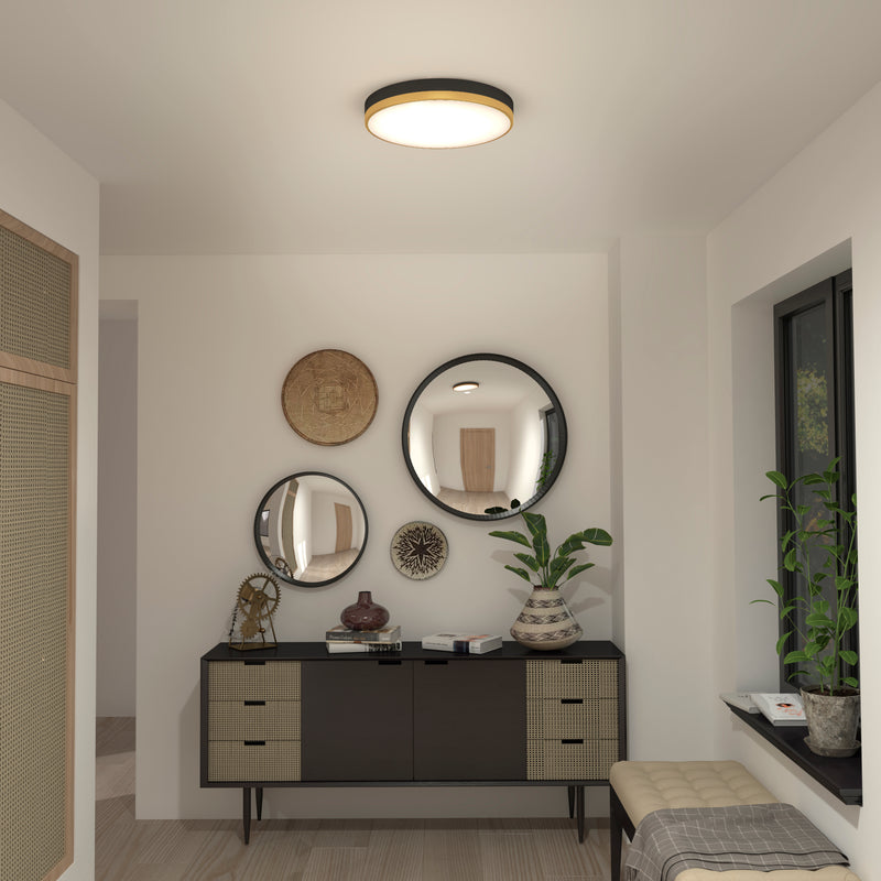 A modern hallway features an Artika Kent LED flush mount light on the ceiling, a black and beige sideboard below three round mirrors, decorative vases and plants, a woven wall basket, and a window with a cushioned bench on the right.