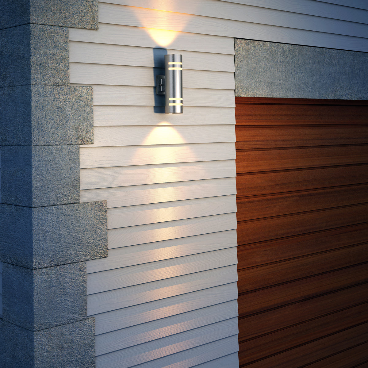 A modern outdoor wall light fixture illuminates the white siding and gray stone trim next to a closed wooden garage door. The light casts a soft glow both upward and downward.