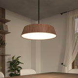 A modern dining room with a wooden table, beige chairs, a large wood-trimmed pendant light, potted plant, shelves with decor, and sheer curtains over a window letting in natural light.