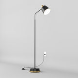 A modern black floor lamp with a gold accent stands on a light gray floor, plugged into a wall outlet. The lamp is turned on, casting light downward from its angled shade.