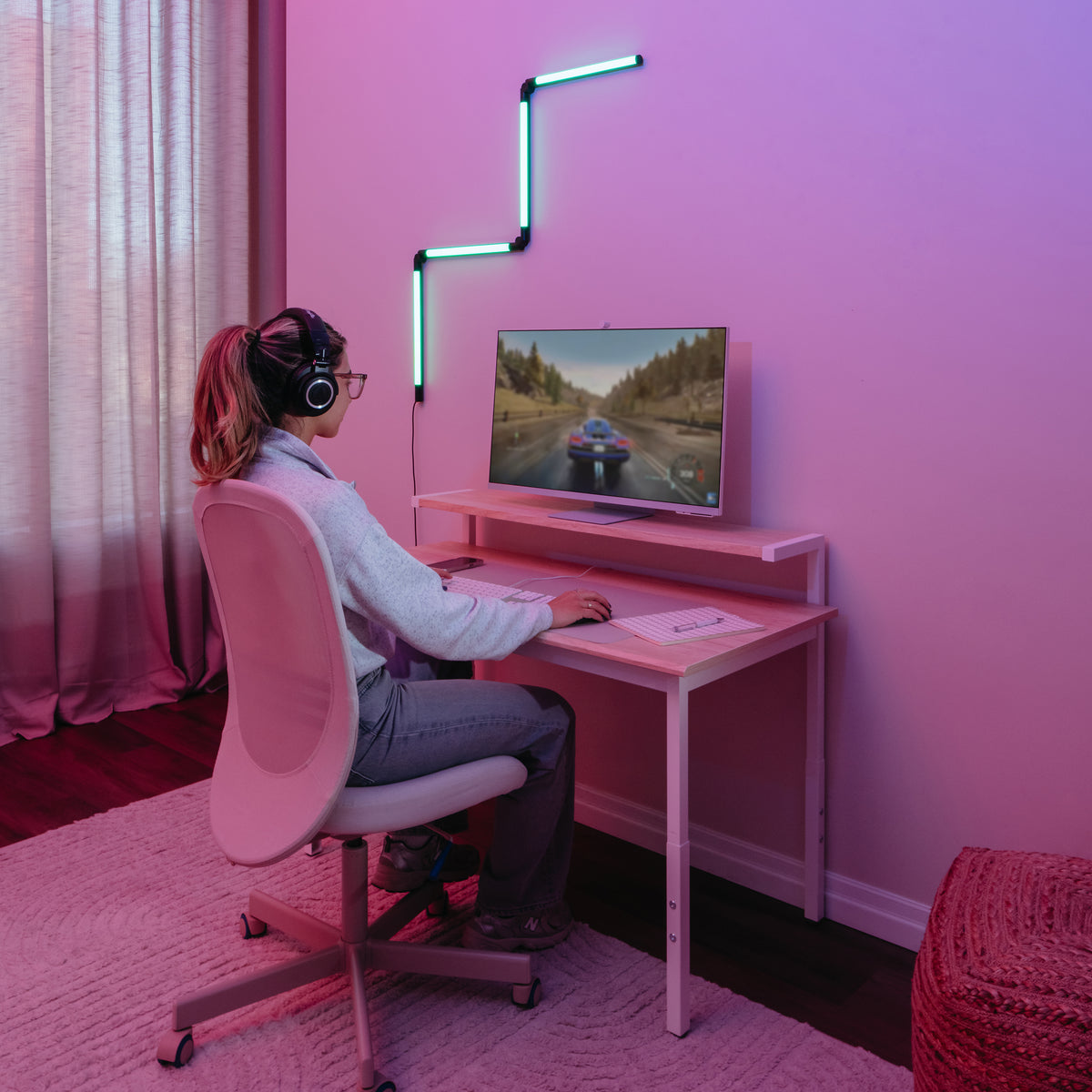 A person wearing headphones sits at a desk playing a car racing game on a monitor. The room is lit with pink and blue neon lights, and the setup includes a white chair, keyboard, and mouse.