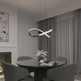 A modern dining room with a round black table, four dark green velvet chairs, and a sculptural LED pendant light above. A fireplace, abstract wall art, and sheer grey curtains complete the elegant, neutral-toned decor.