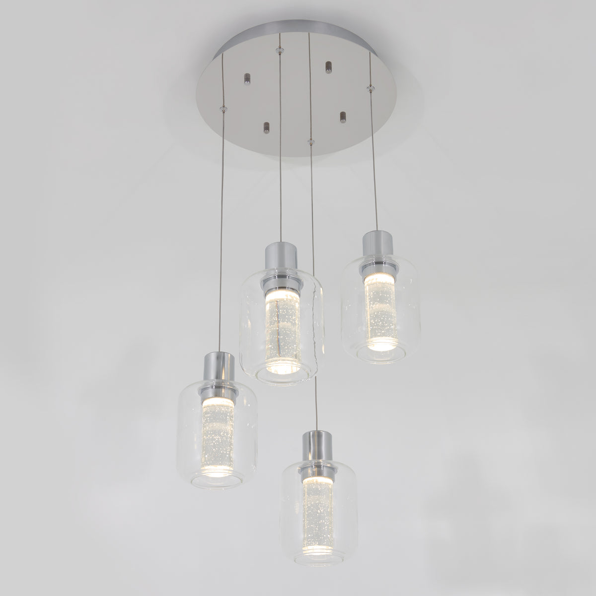 A modern ceiling light fixture with four cylindrical glass pendant lights, each suspended at different heights from a round silver base, emitting a soft white glow.
