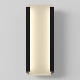 A modern wall sconce with a rectangular, vertical design featuring black side panels and a soft, warm light glowing from the center against a light gray wall.