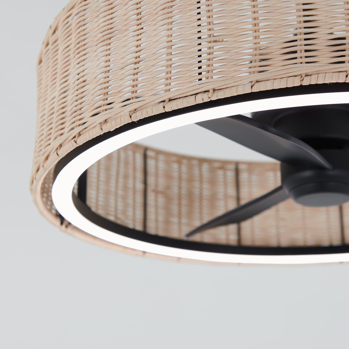 A close-up view of a ceiling fan with black blades, surrounded by a circular LED light and a woven rattan shade, set against a light gray background.
