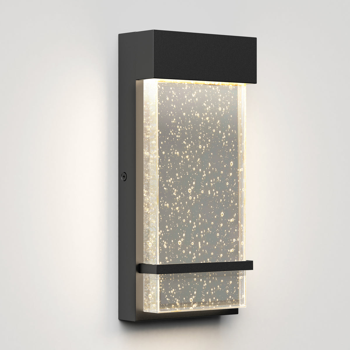 A modern wall sconce with a rectangular black frame and a clear glass panel featuring air bubbles, illuminated with soft, warm white light, mounted on a light-colored wall.