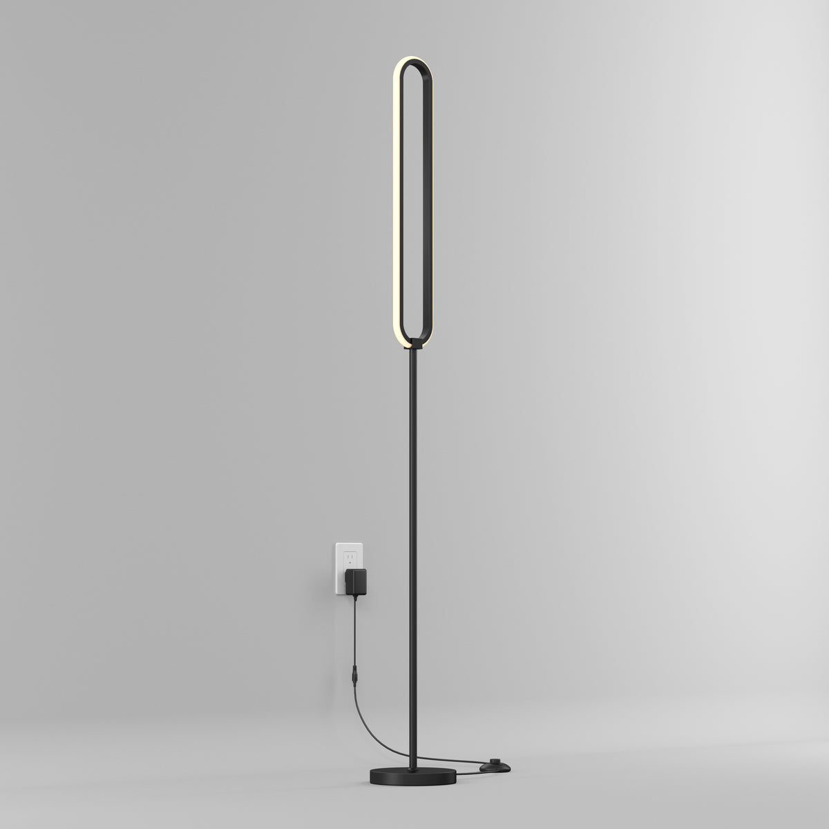 A modern, minimalist floor lamp with an oval-shaped, illuminated LED frame stands on a black base against a plain white wall, plugged into an electrical outlet with a visible black power cord.