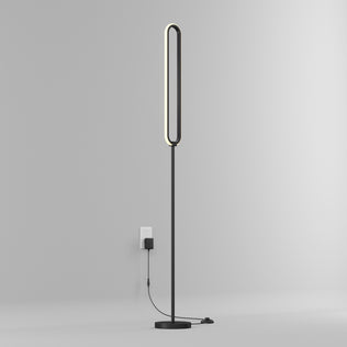 The Artika Leni dimmable LED modern floor lamp, featuring a sleek minimalist design and elongated oval light, stands against a plain gray wall and is plugged into a wall outlet.