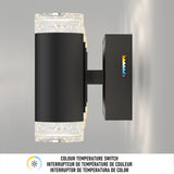 A modern black wall sconce with illuminated textured glass ends is mounted on a wall. A color temperature switch with five settings is visible on the side. Text below describes the color temperature switch in four languages.