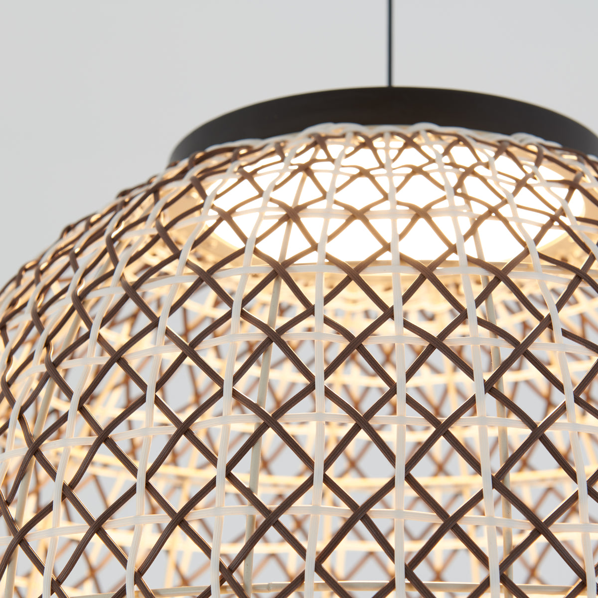 Close-up of a woven rattan lampshade with a crisscross pattern, illuminated from within and hanging from the ceiling by a black cord, against a light background.