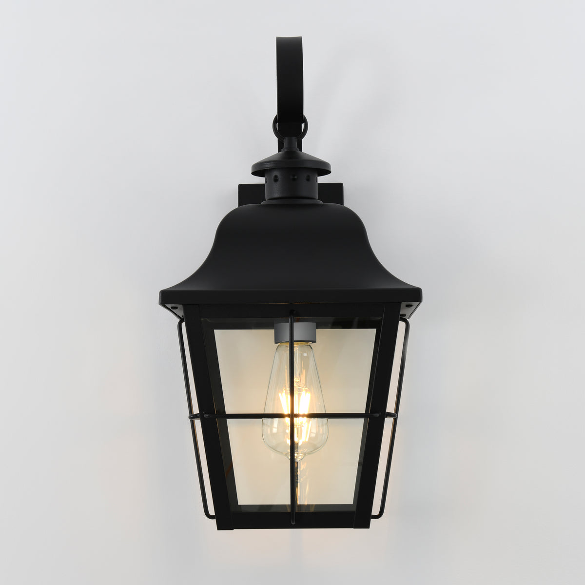 A black metal outdoor wall lantern with a clear glass enclosure, mounted on a white wall, and featuring an exposed glowing Edison-style bulb inside.
